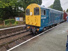 a blue and yellow train traveling down tracks next to a loading platform with people looking on