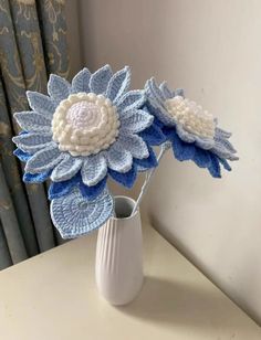 two crocheted flowers in a white vase on a table next to a curtain