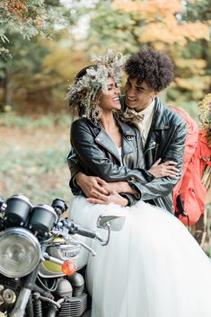 a man and woman sitting on top of a motorcycle in front of trees with leaves