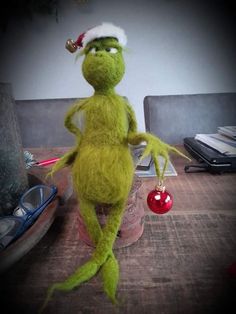 a green stuffed animal wearing a santa hat and holding a red ornament on top of a wooden table