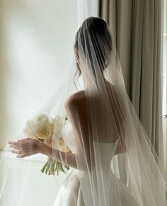 a woman in a wedding dress is holding a bouquet and looking out the window with her veil over her head