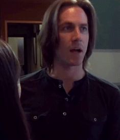 a man with long hair standing in front of a mirror talking to another man who is wearing a black shirt