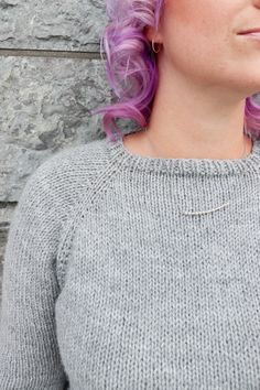 a woman with pink hair wearing a gray sweater and silver earring, standing in front of a stone wall