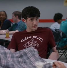 a young man sitting at a table with other people in the background eating and talking