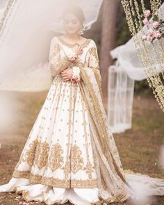 a woman in a white and gold wedding gown standing under an arch with flowers on it