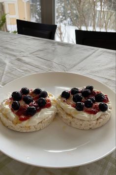 two pastries with blueberries and cream on a white plate next to a window