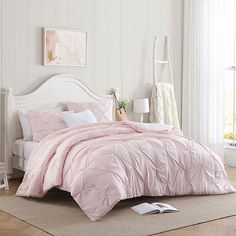 a bed with pink comforter and pillows in a white room next to a window