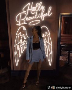 a woman standing in front of a neon sign with angel wings on it's side