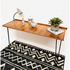 a wooden table with two plants on top and a rug underneath it in front of a white wall