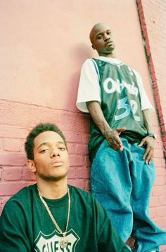 two young men leaning against a pink brick wall, one is wearing a green shirt and the other wears blue jeans