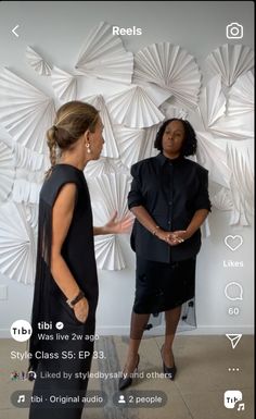 two women standing next to each other in front of a wall with paper sculptures on it