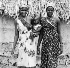 two women and a baby standing in front of a thatched hut with straw roof