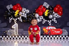 a baby boy sitting on the floor in front of a birthday cake backdrop with cars