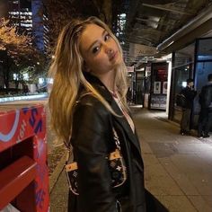 a woman standing next to a red bench on a city street at night with her hair blowing in the wind