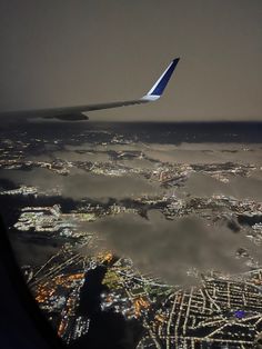 the wing of an airplane flying over a city at night with lights and clouds in the foreground