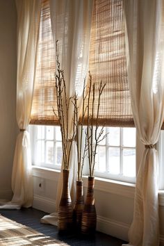 three tall vases with branches in front of a window covered by sheer drapes