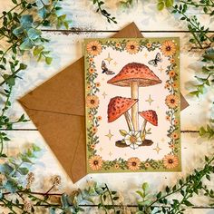 a card with flowers and mushrooms on it sitting on top of a wooden table next to a brown envelope