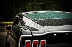 the front end of a black muscle car covered in rain and condensation with trees in the background