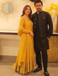 a man and woman standing next to each other in front of a wall with yellow flowers