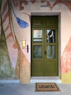 a green door is on the side of a pink building with trees painted on it