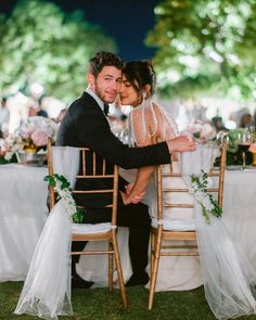 a bride and groom sitting at a table with their hands on the back of chairs