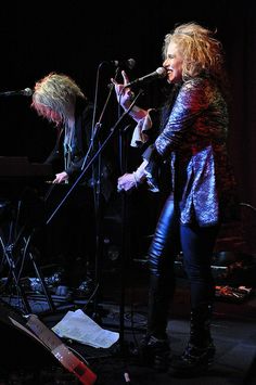 two women on stage singing into microphones with music instruments in front of them,