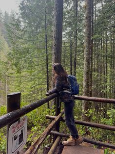 a woman with a backpack standing on a wooden bridge in the woods looking at trees