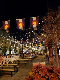 people are sitting at tables in an outdoor area with lights strung over the buildings and trees