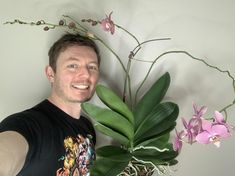 a man standing next to a plant with purple flowers on it and smiling at the camera