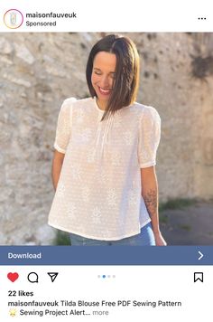 a woman wearing a white blouse and jeans standing in front of a stone wall with her hand on her hip