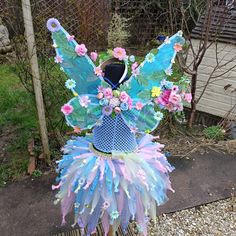 a blue and pink fairy costume with flowers on it's head, sitting in front of a fence