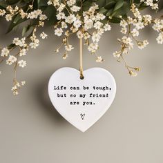 a white heart hanging from a branch with flowers in the background and a quote written on it that says, life can be tough but so my friends are you