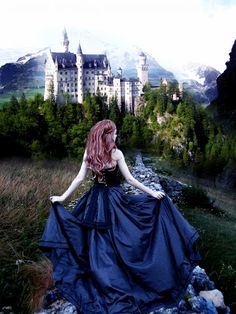 a woman in a blue dress is sitting on rocks near a castle with mountains behind her