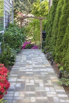 a brick walkway surrounded by flowers and greenery in front of a house with a pergolated arbor