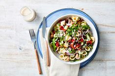 a white bowl filled with pasta salad next to a fork and spoon on top of a blue plate