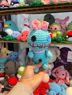 a hand holding a small crocheted toy in front of a shelf full of stuffed animals