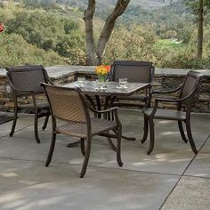 an outdoor dining table with four chairs and a vase of flowers on the table in front of a stone wall