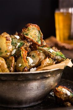 a metal bowl filled with potatoes covered in cheese and herbs next to a glass of beer