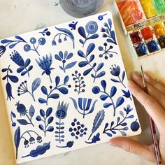 a hand holding a paintbrush next to a blue and white flower design on a tile