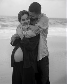 a pregnant woman hugging her husband on the beach in front of the ocean, black and white photograph