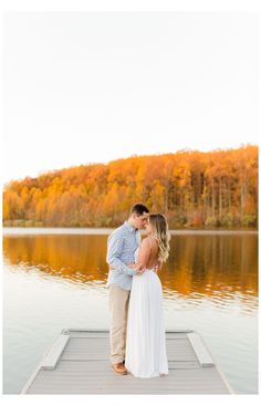 an engaged couple standing on a dock in front of a lake surrounded by fall foliage