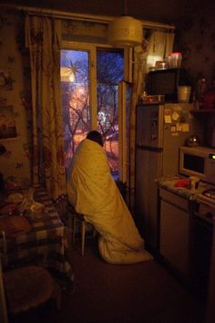 a person wrapped in a blanket sitting at a kitchen window looking out onto the yard