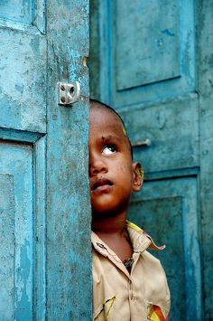 a young boy peeks his head out from behind a blue door, looking up at the sky