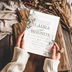 a person holding up a wedding card on top of a wooden table next to flowers