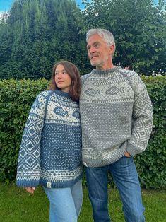 an older man and woman standing next to each other in front of some bushes, wearing sweaters