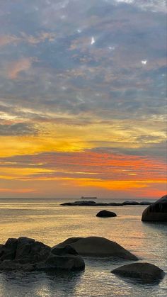 an orange and yellow sunset over the ocean with rocks in the foreground, and small island in the distance