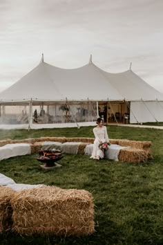 a woman sitting on hay bales in the middle of a field next to a large white tent