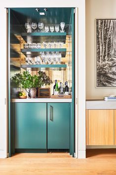 an open cabinet with wine glasses and plants on it in a living room next to a painting