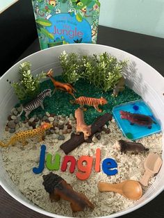 a white bowl filled with sand and plastic animals on top of a table next to a book