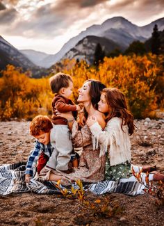 a woman sitting on the ground with her two children in front of mountains and trees